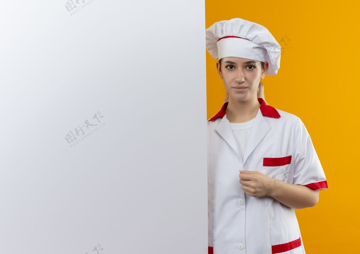 站立自信的年轻漂亮厨师穿着厨师制服站在白墙后面 手放在她隔离在橙色墙上的制服上 留有复印空间烹饪自信手