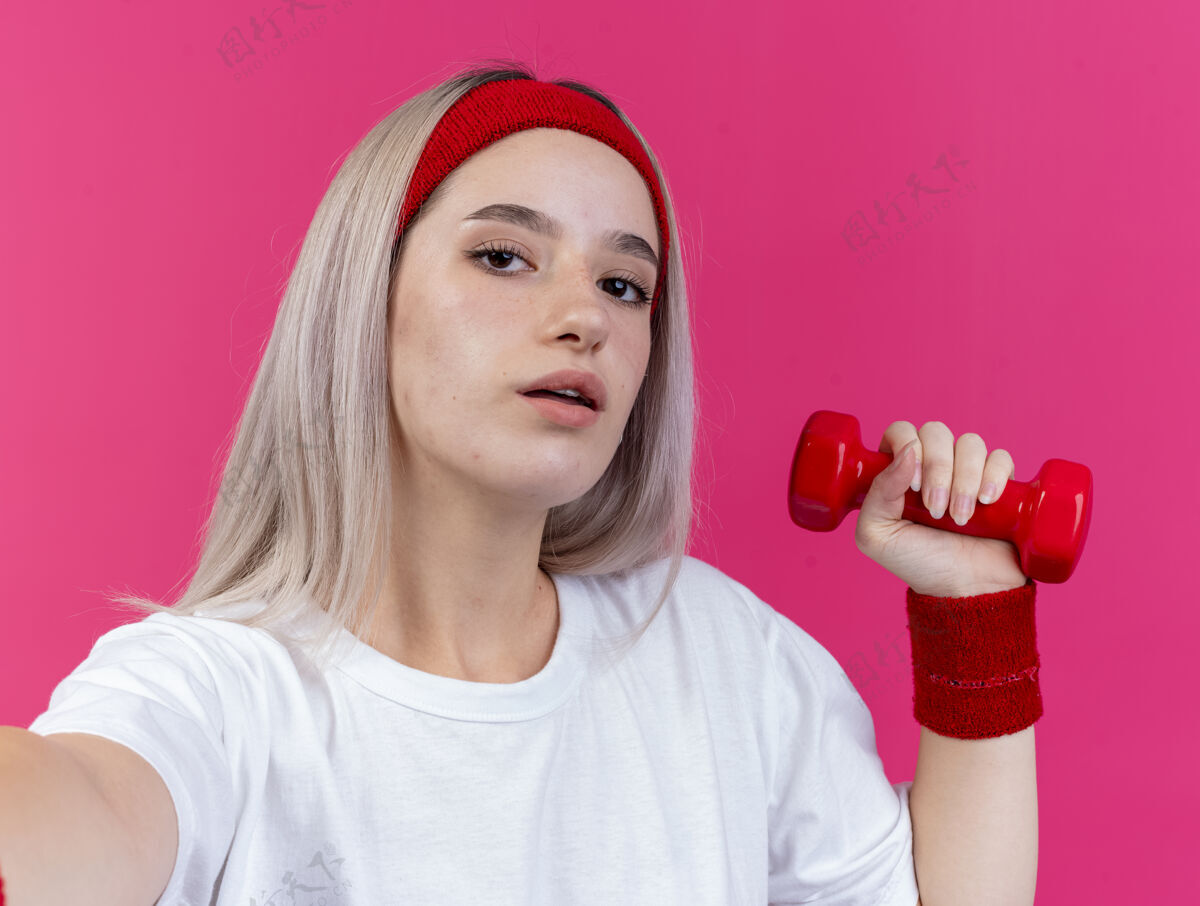 粉红自信的年轻白人运动女孩戴着背带 戴着头带和腕带 举着哑铃看着镜头信心哑铃背带