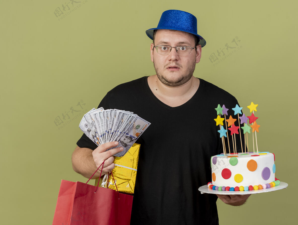 穿震惊的成年斯拉夫男子戴着眼镜 戴着蓝色派对帽 手里拿着钱礼盒 纸购物袋和生日蛋糕蓝色购物眼镜