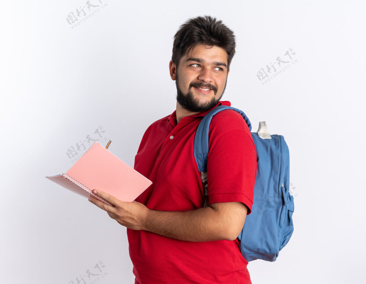 红色年轻的留着胡子的学生 穿着红色马球衫 背着书包 站在笔记本上写着自信的微笑胡须学生写作
