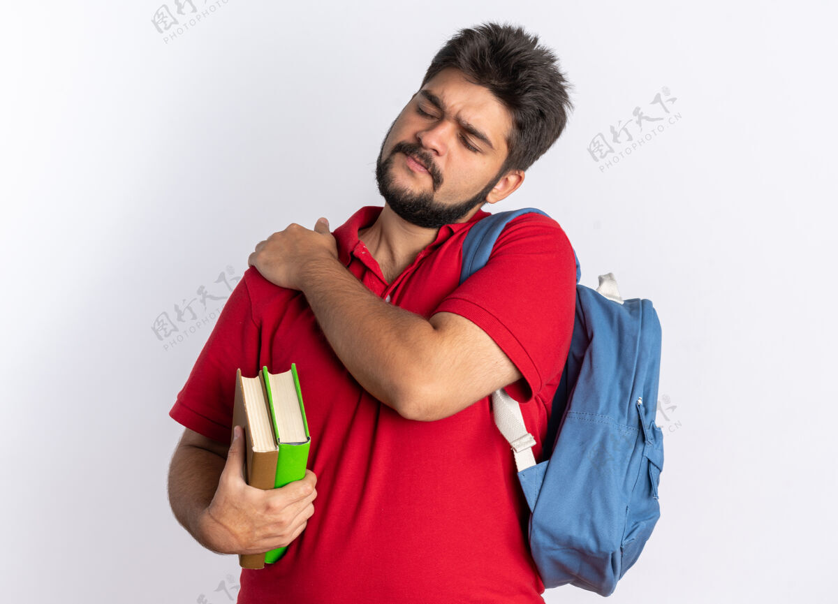 抱着年轻的留着胡子的学生 穿着红色马球衫 背着背包 拿着笔记本 看起来不舒服 摸着肩膀 站着感觉很痛触摸肩膀背包