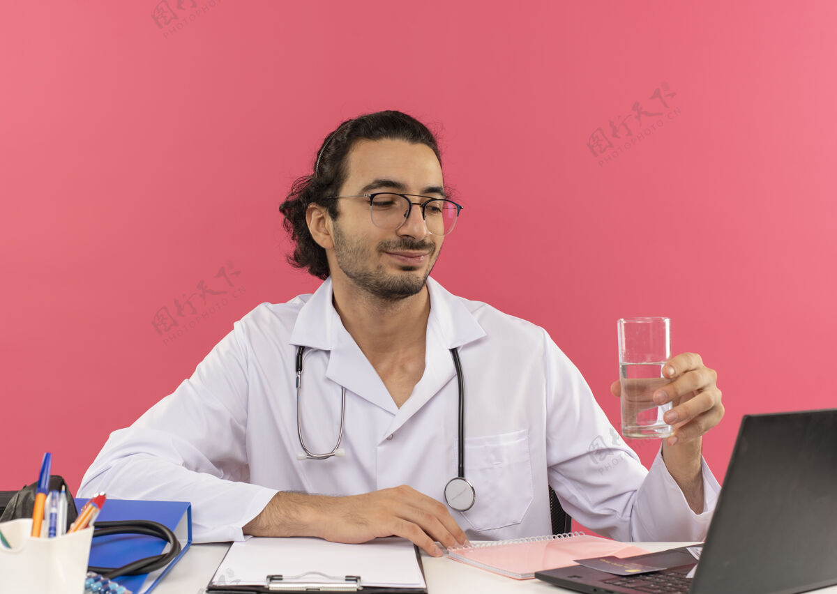 复印年轻的男医生戴着医用眼镜 穿着医用长袍 听诊器坐在桌边 很高兴坐着的桌子粉色