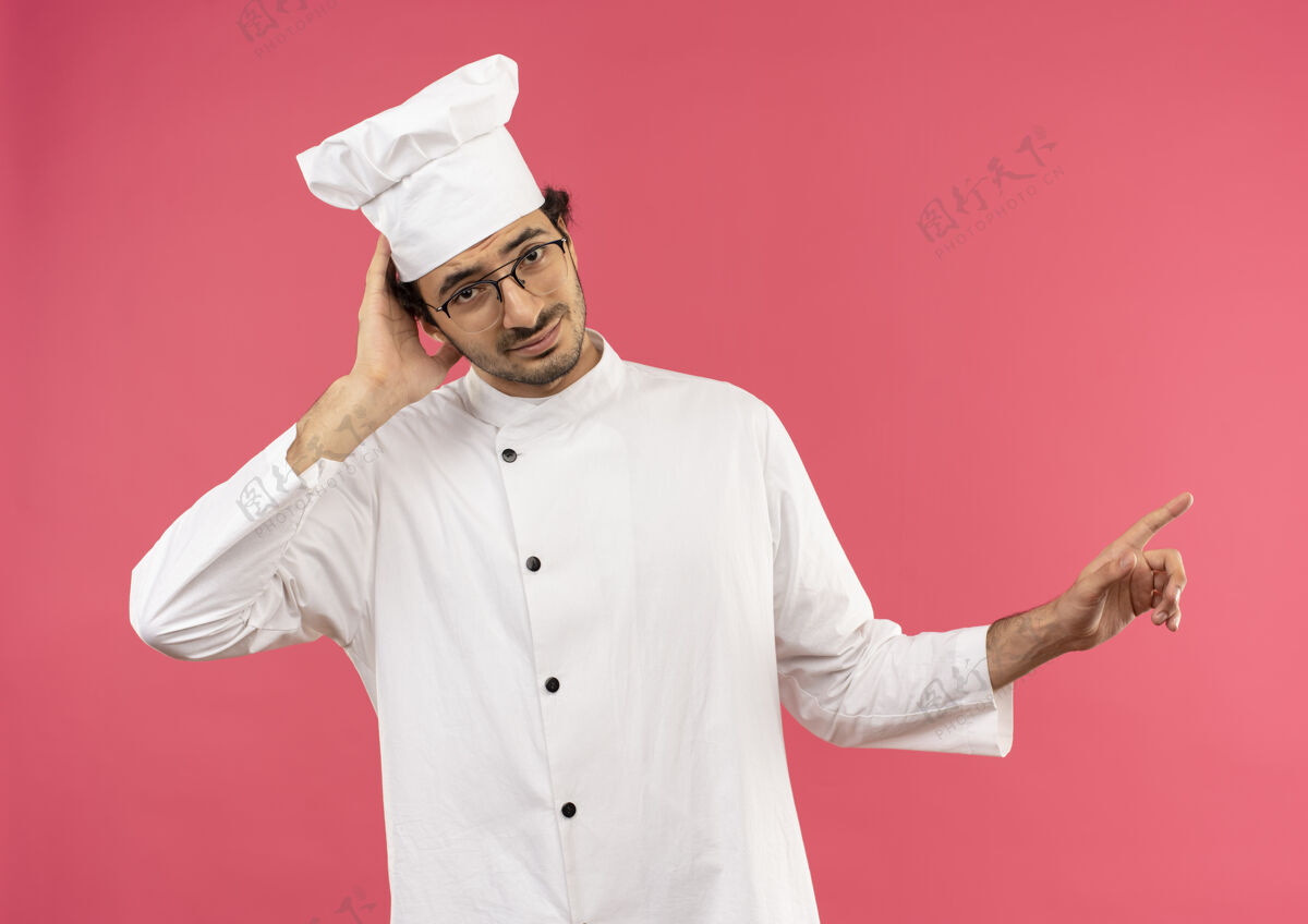 男看上去很困惑的年轻男厨师穿着厨师制服 戴着眼镜 手放在头上 指着旁边烹饪制服手