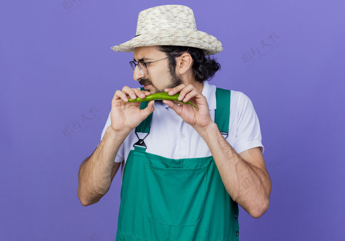 绿色年轻的留着胡须的园丁 穿着连体衣 戴着帽子 脸上露出绿色的辣椒 站在蓝色的背景上 看着一旁扭动着男人胡子帽子