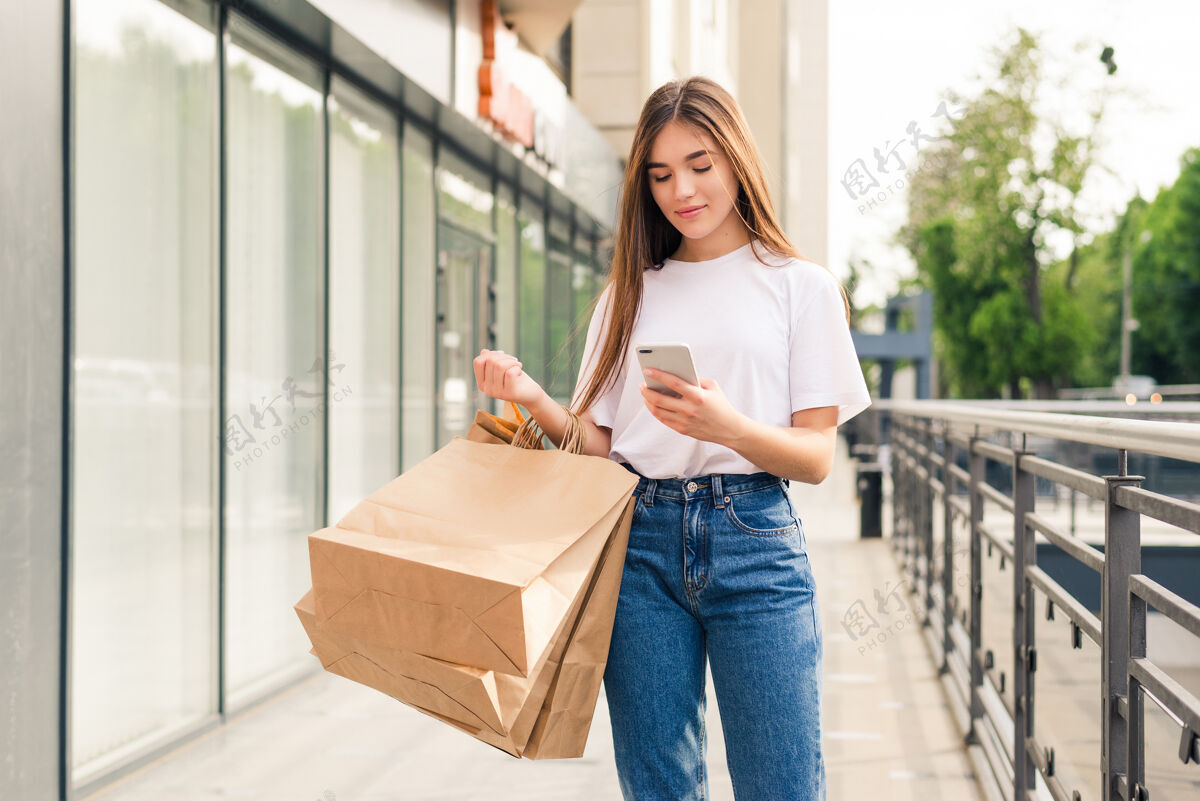 爱好与朋友分享好消息美丽的年轻微笑的女人拿着购物袋和手机站在户外的特写镜头移动手消费主义