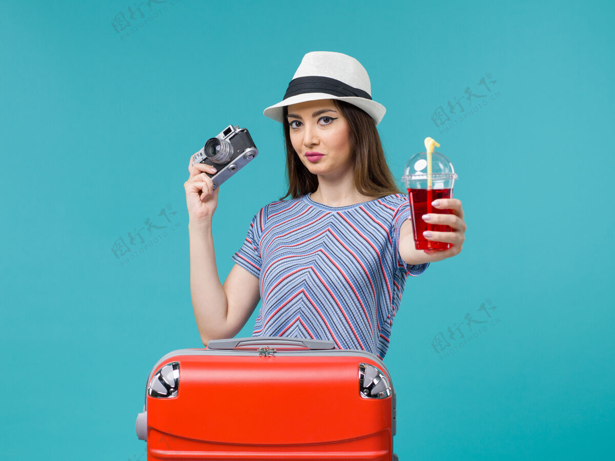 旅程前景：度假中的女人拿着果汁和相机在蓝色的背景上海上避暑旅行飞机旅行果汁帽子举行