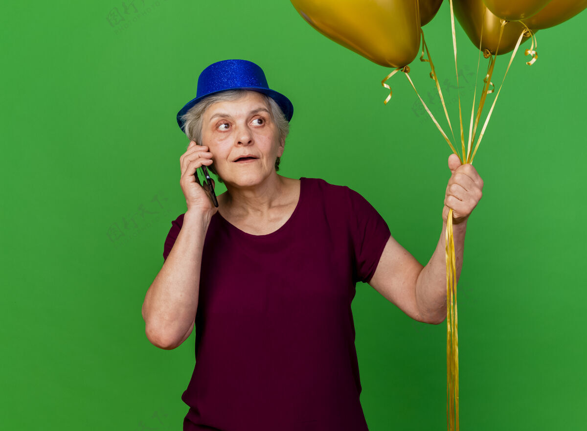 派对戴着派对帽的惊讶的老妇人手持氦气球 一边打电话一边看着绿色气球惊喜电话