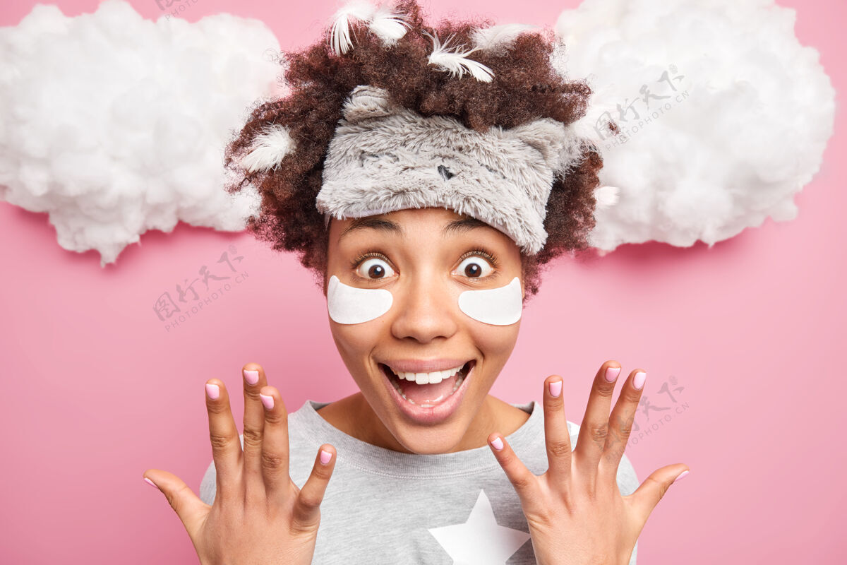 睡衣积极兴奋的卷发女人举手微笑开心有惊喜表情羽毛扎在头发上戴着睡衣睡衣被惊喜的好消息隔离在粉色的墙上放松休闲羽毛