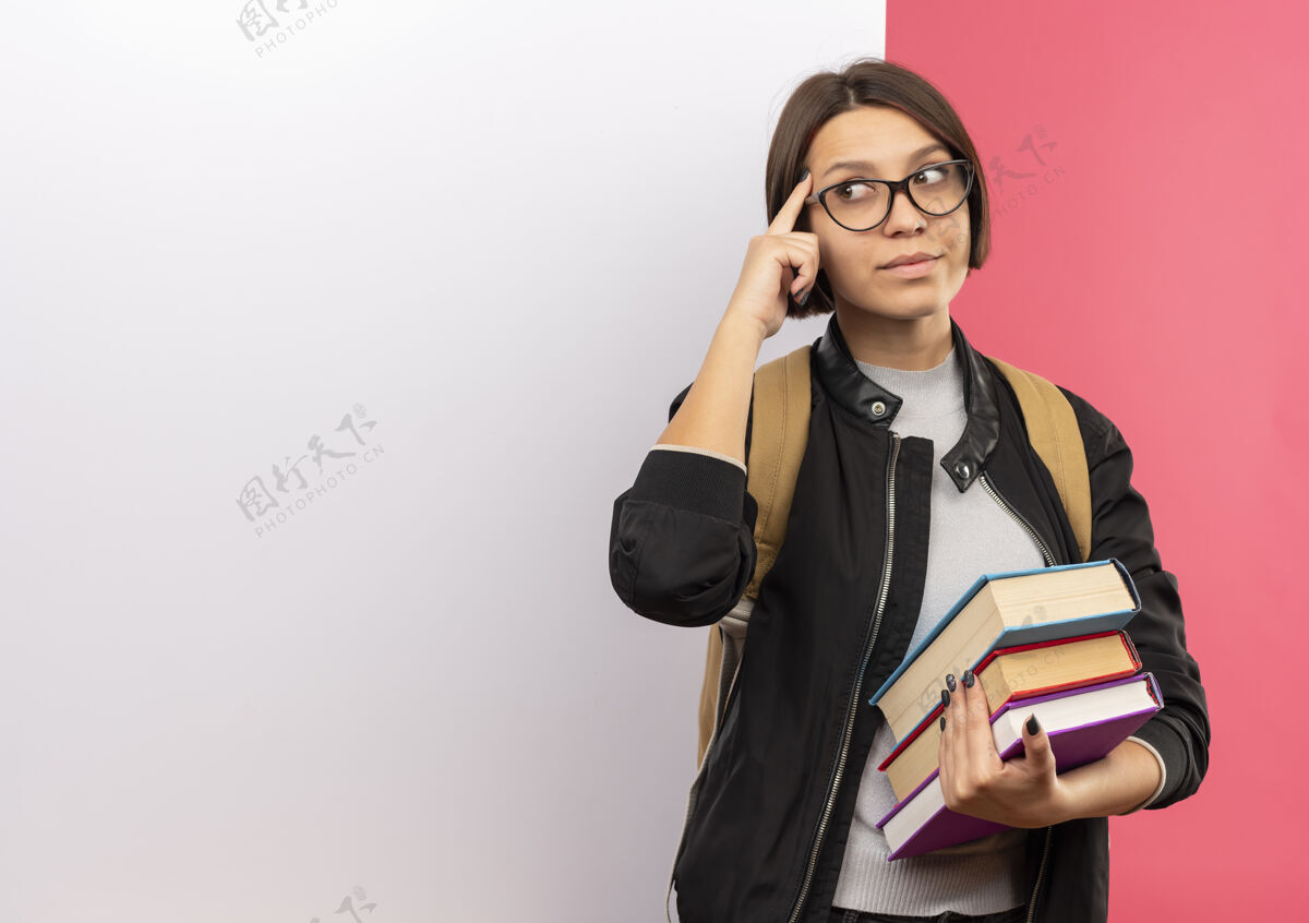 姿势体贴的年轻女学生戴着眼镜 背着书包 站在白色的墙前 手指放在粉红色的墙上孤立的太阳穴上 看着侧面人眼镜表情