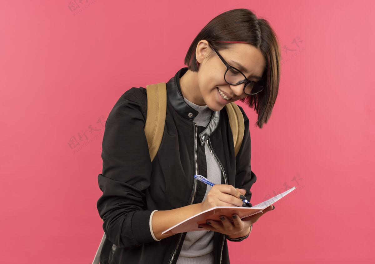 表情微笑的年轻女学生戴着眼镜 背着书包 在粉红色墙上的便笺簿上写着什么人包脸