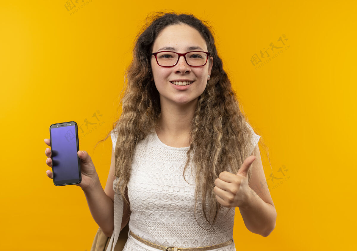 拇指微笑着的年轻漂亮的女学生戴着眼镜 背着书包 拿着手机 大拇指向上竖起 孤零零地站在黄色的墙上感觉表情人