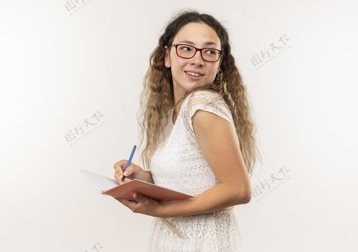 感觉面带微笑的年轻漂亮的女学生戴着眼镜 背着书包站在侧视图上 看着身后的笔迹 把笔记本上的笔孤立在白色的墙上穿女生人