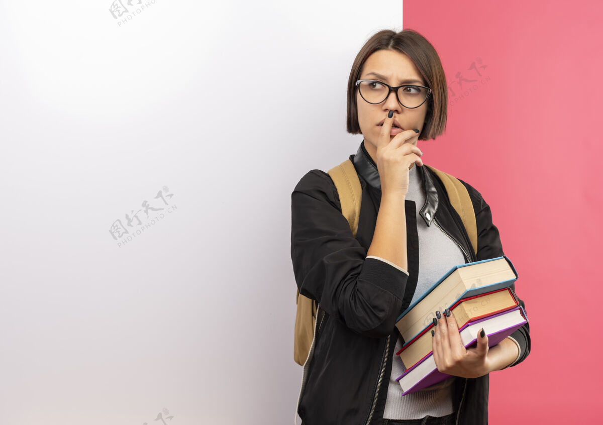 人体贴的年轻女学生戴着眼镜 背着书包 站在白色的墙前 抚摸着下巴 看着粉红色墙上孤立的一面感觉壁板前面