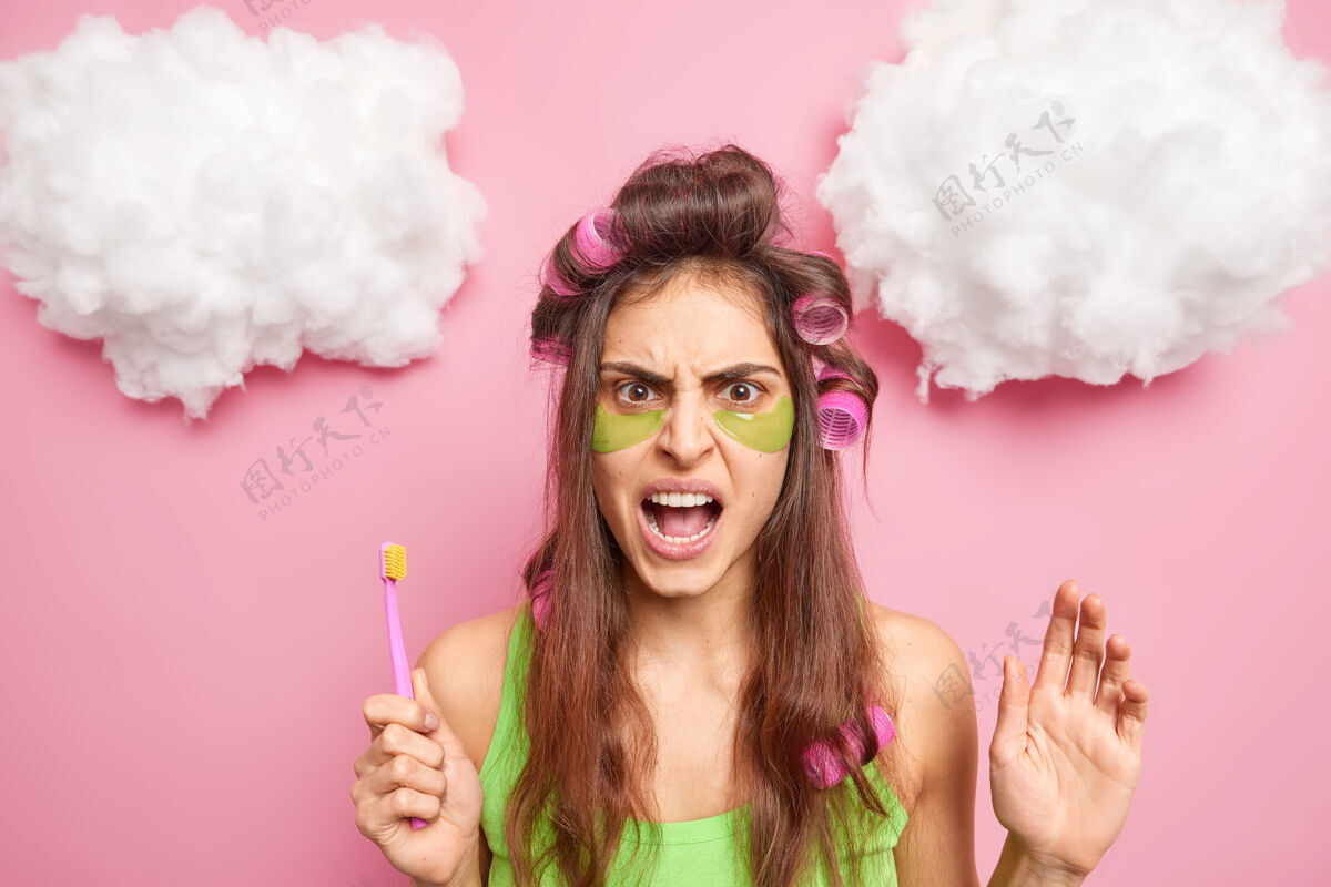 成人生气的黑发女人在眼睛下面涂上卷发器胶原蛋白贴片来减少皱纹和细纹拿着牙刷把每天的口腔卫生习惯隔离在粉色的工作室墙上新鲜愤怒女人