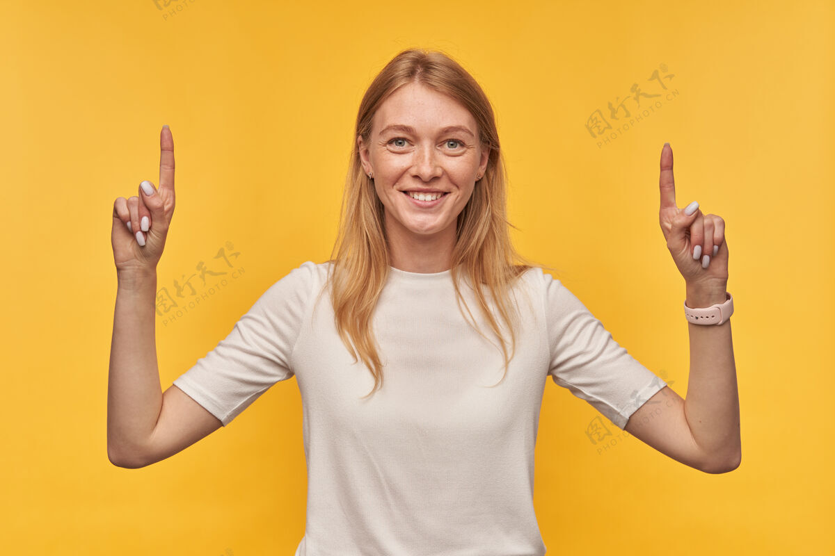 魅力一个满脸笑容 长着雀斑 穿着白色T恤的漂亮女人站着 用两只手指指着黄色的双手站立欧式手指