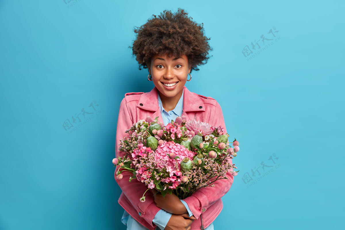 微笑室内拍摄的积极的年轻女子拥抱大束鲜花微笑愉快地穿着粉红色夹克隔离在蓝色的墙壁事件卷曲满意
