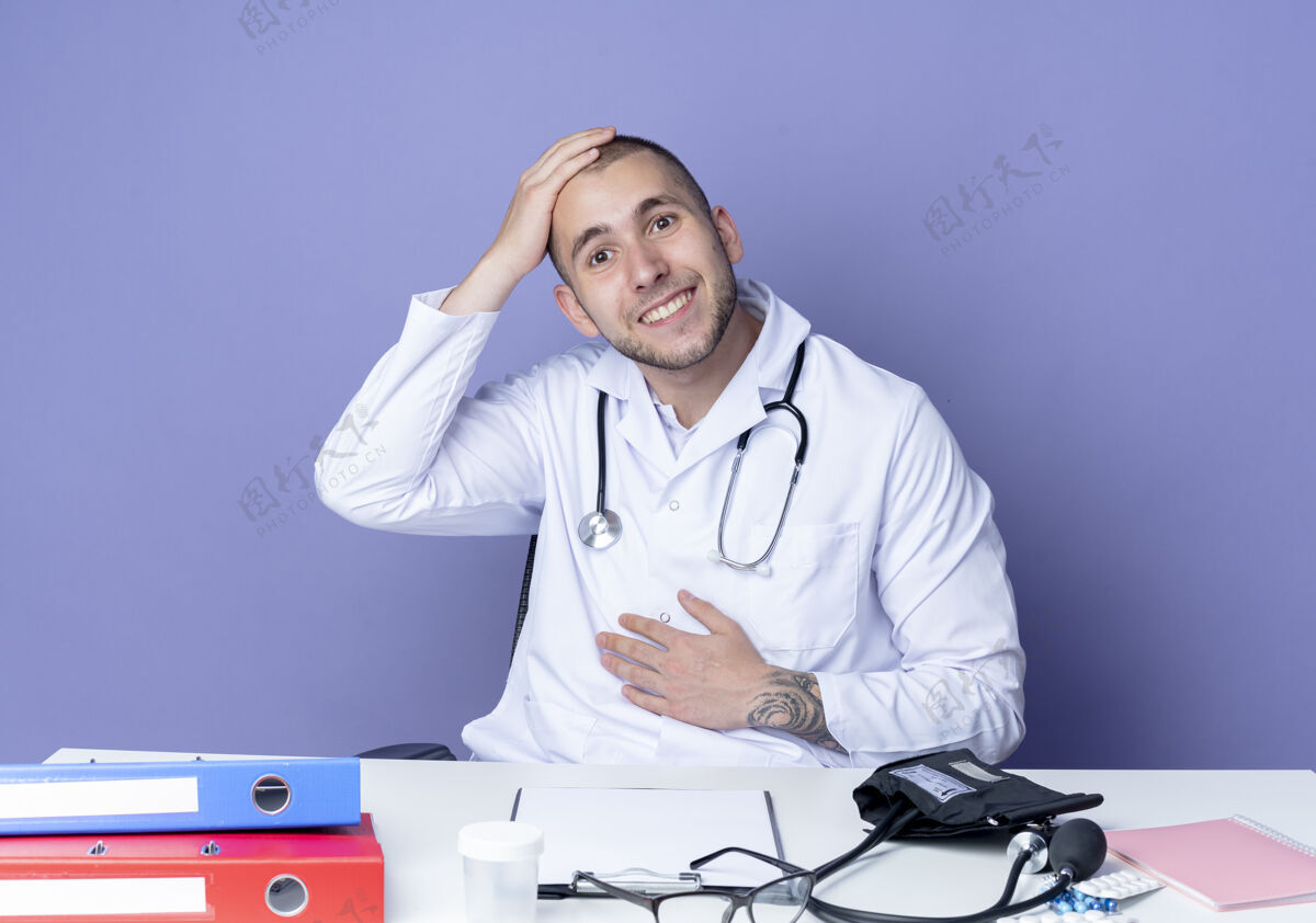 男性面带微笑的年轻男医生穿着医用长袍和听诊器坐在办公桌旁 手放在肚子上 头孤立在紫色的墙上头表情长袍