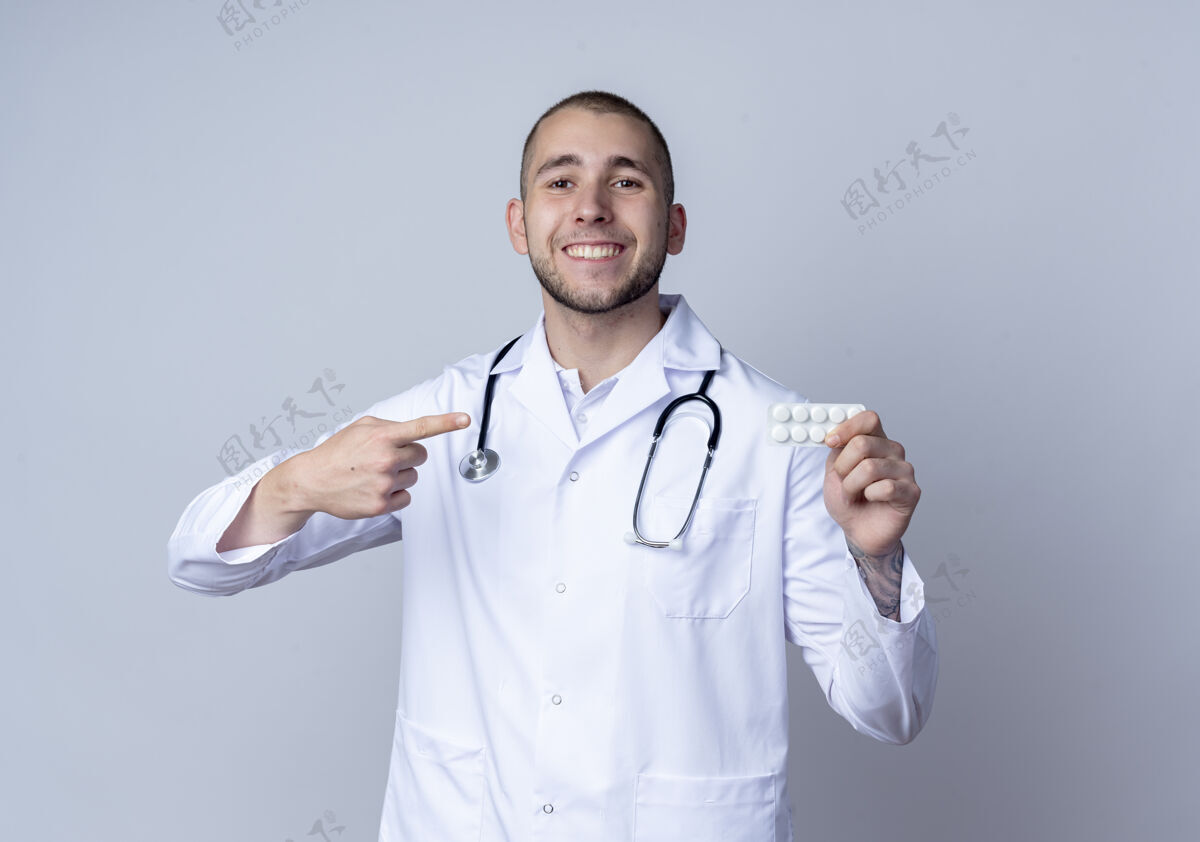 药片面带微笑的年轻男医生 穿着医用长袍 脖子上戴着听诊器 拿着并指着隔离在白墙上的一包药片白人包感情