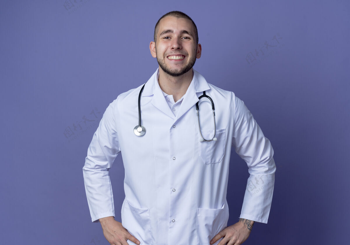 表情面带微笑的年轻男医生 穿着医用长袍 手放在紫色墙上隔离的腰上 戴着听诊器人手微笑