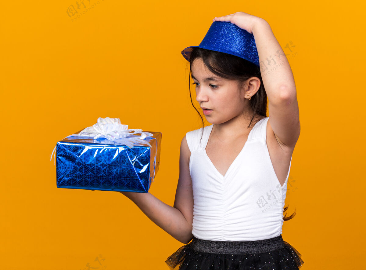 惊讶惊讶的年轻白人女孩戴着蓝色派对帽 手放在帽子上 看着孤立在橙色墙上的礼品盒 还有复制空间帽子手女孩
