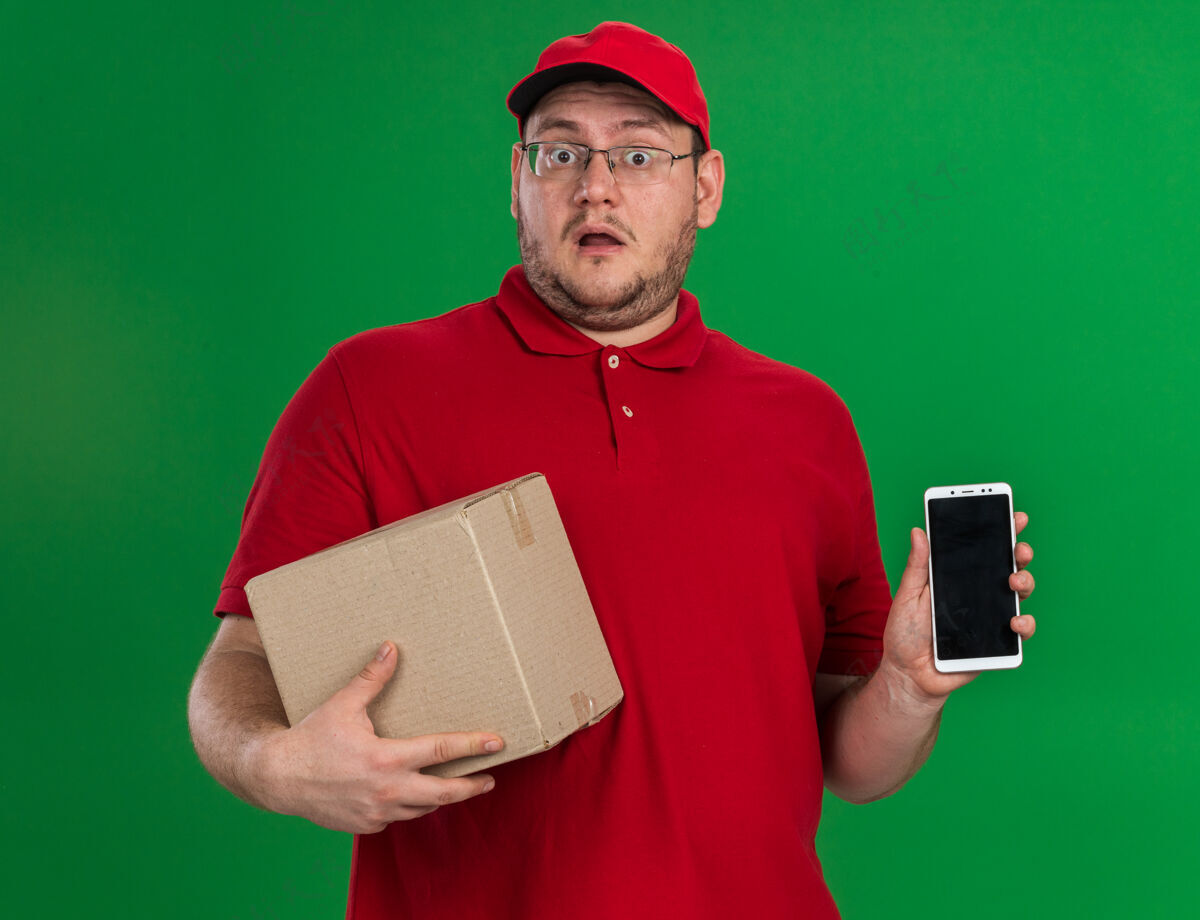 送货员震惊超重的年轻送货员 戴着眼镜 拿着纸板箱和电话 隔离在绿墙上 留着复印空间手持光学眼镜