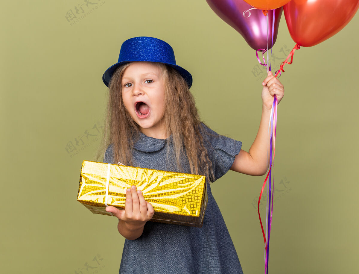 氦惊讶的金发小女孩 戴着蓝色派对帽 手里拿着氦气球和礼盒 隔离在橄榄绿的墙上 还有复印空间小生日橄榄