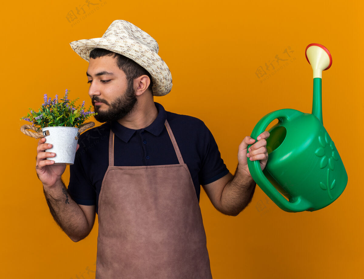 浇水高兴的年轻白人男性园丁戴着园艺帽拿着浇水罐在花盆里嗅花香隔离在橙色的墙上复制空间穿隔离请