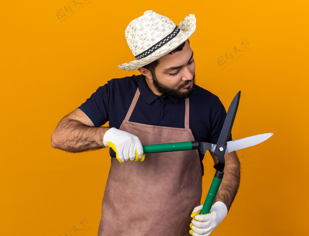 手套自信的年轻白人男性园丁戴着园艺帽子和手套拿着园艺剪刀看着隔离在橙色墙上的复制空间年轻剪刀花园