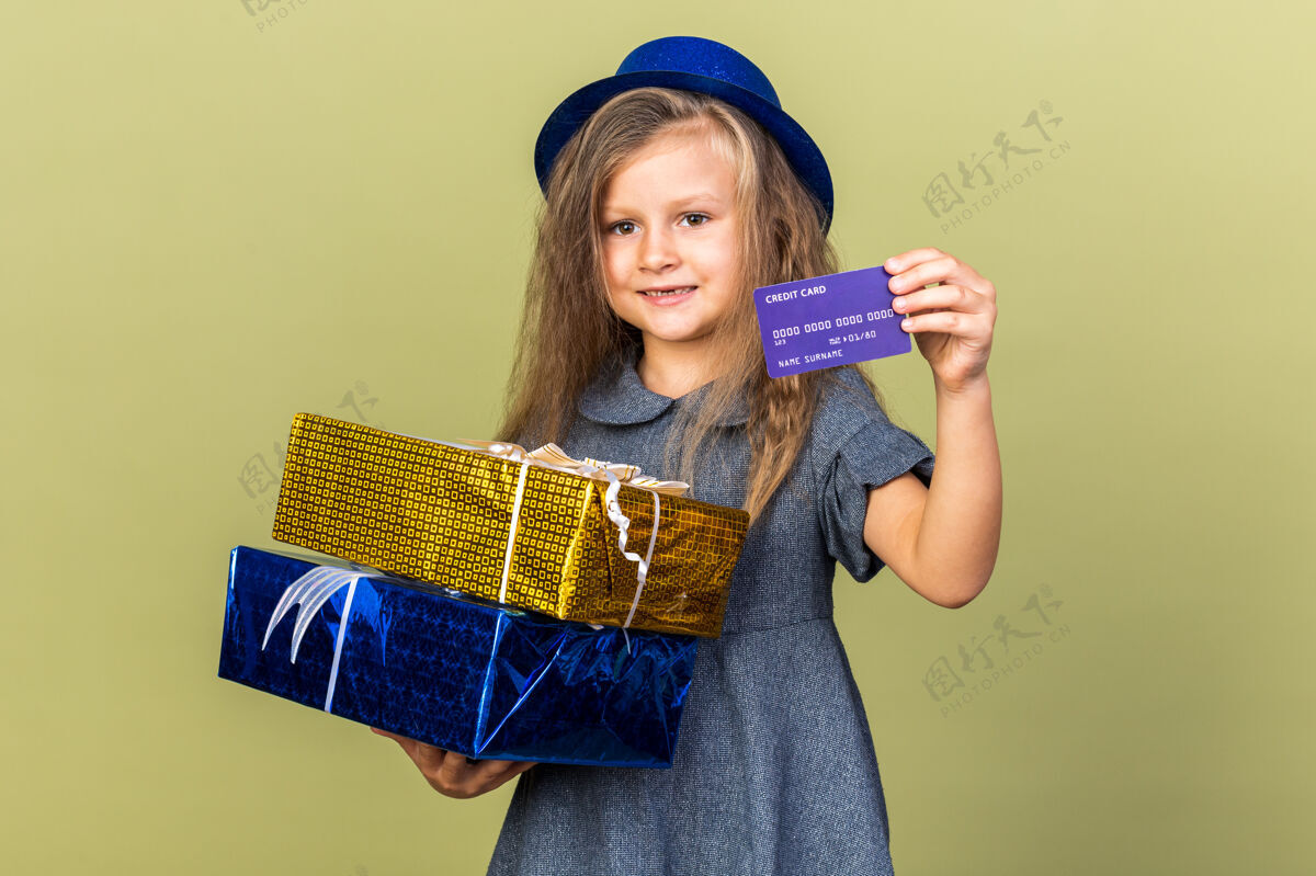 信用卡微笑的金发小女孩 戴着蓝色派对帽 拿着礼品盒和信用卡 隔离在橄榄绿的墙上 留着复印空间派对帽子持有