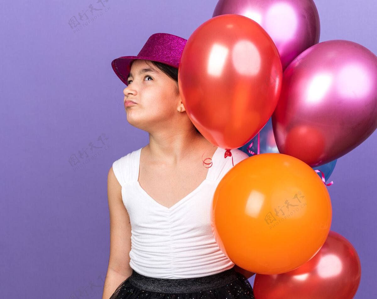 孤立戴着紫色派对帽 手持氦气球 孤零零地仰望着紫色墙壁 留有复制空间的年轻白人女孩女孩不愉快年轻