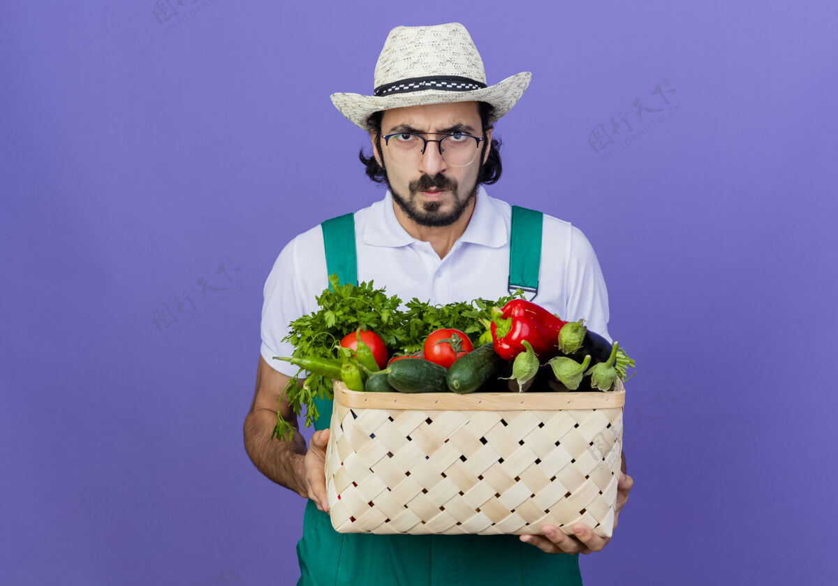 连身衣年轻的留着胡须的园丁 穿着连体衣 戴着帽子 手里拿着装满蔬菜的箱子 站在蓝色的墙上 严肃地皱着眉头看着前方皱眉站着表情
