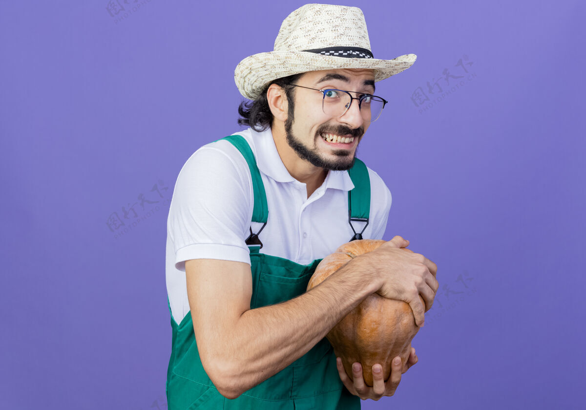 人年轻的留着胡子的园丁 穿着连体衣 戴着帽子 手里拿着南瓜 站在蓝色的墙上 狡猾地微笑着看着前面花园抱着胡须