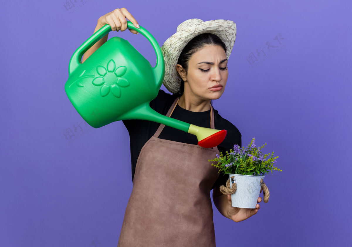 花园年轻漂亮的女园丁围着围裙 戴着帽子 手里拿着浇水罐 站在蓝色的墙上浇灌盆栽植物漂亮衣服人