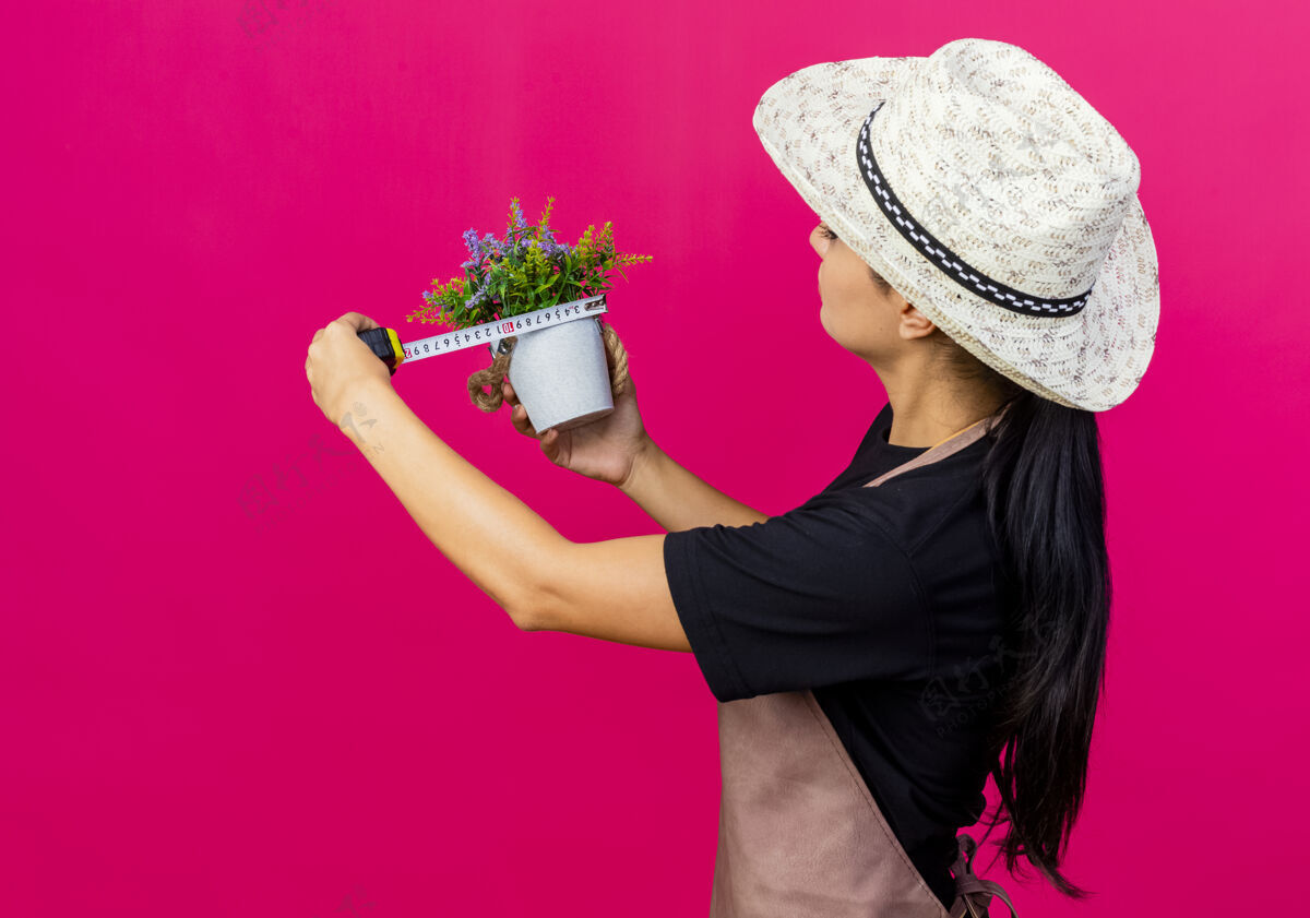 人年轻漂亮的女园丁 围着围裙 戴着帽子 手里拿着盆栽和卷尺 严肃的脸站在粉红色的墙上严肃感觉站