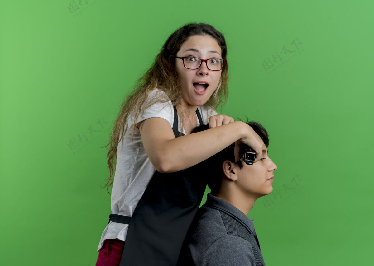 女人年轻的专业美发师女人围着围裙用男顾客的剪子剪头发 站在绿墙上看着眼前大吃一惊姿势围裙表情