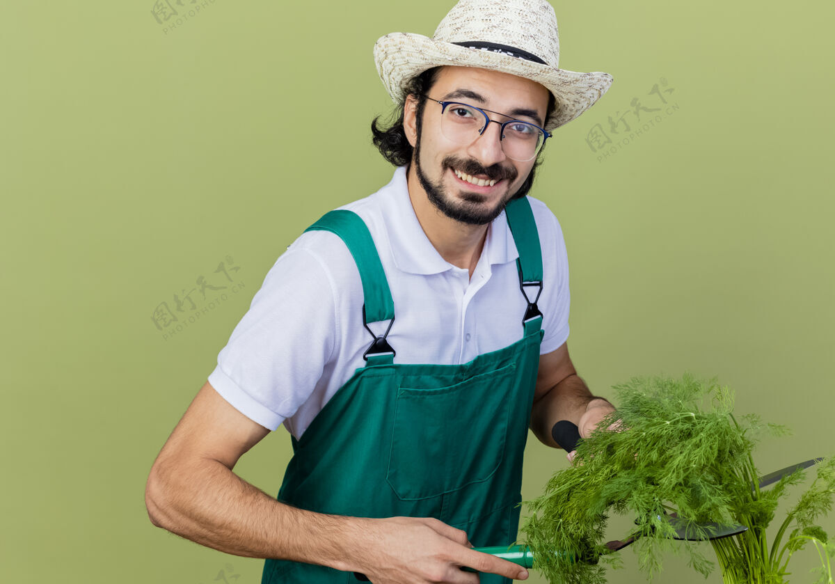 脸年轻的留着胡须的园丁 穿着连体衣 戴着帽子 手里拿着新鲜的草药 站在浅绿色的墙上 面带微笑地看着前面新鲜抱着人