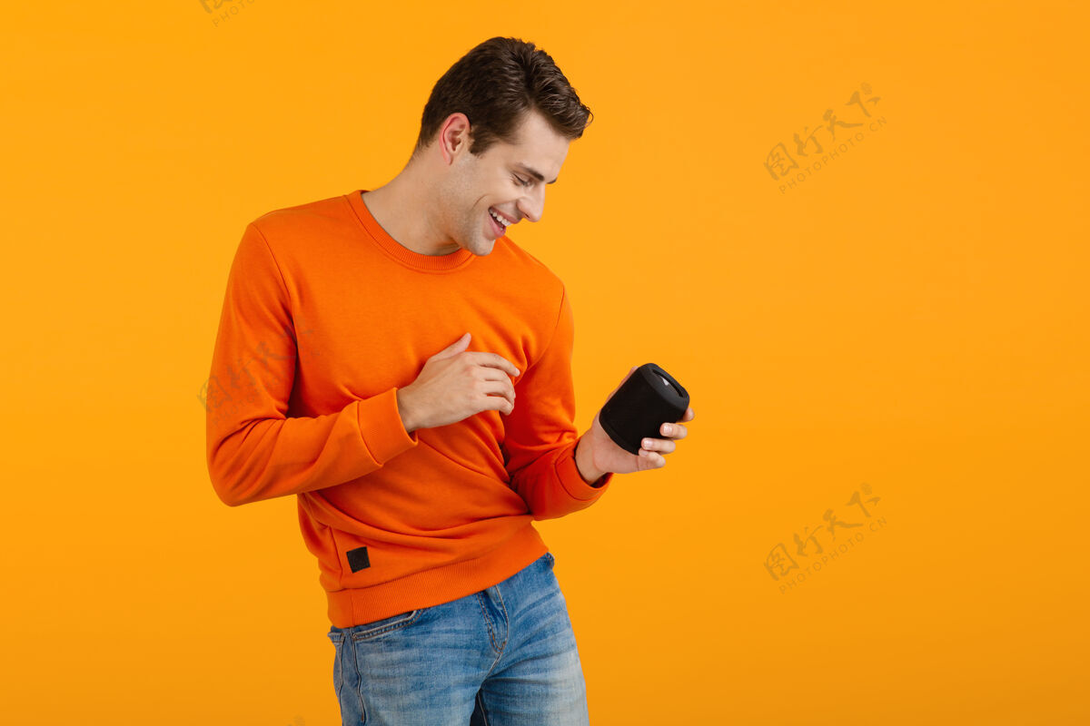 时尚穿着橙色毛衣的时髦微笑的年轻人拿着无线扬声器快乐地听音乐享受橙色的乐趣扬声器男性搞笑