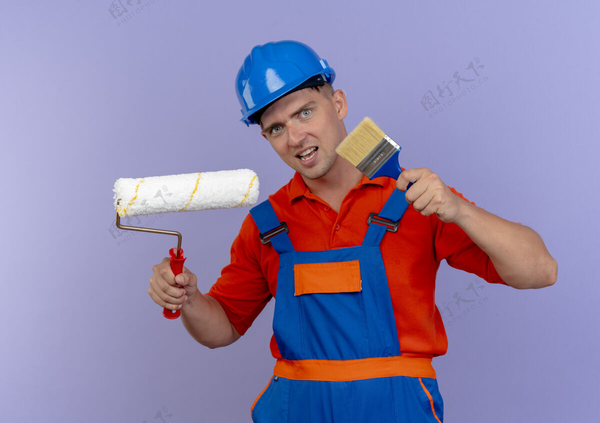 建筑工人高兴的年轻男性建筑工人穿着制服和安全帽举行油漆辊和油漆刷制服安全请