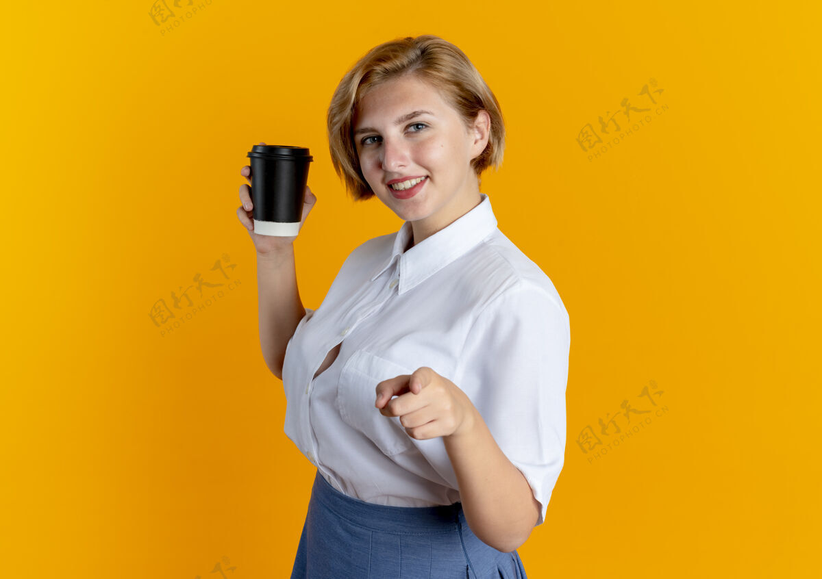 杯子年轻的微笑的金发俄罗斯女孩站在一边拿着咖啡杯 指着镜头背景俄罗斯微笑