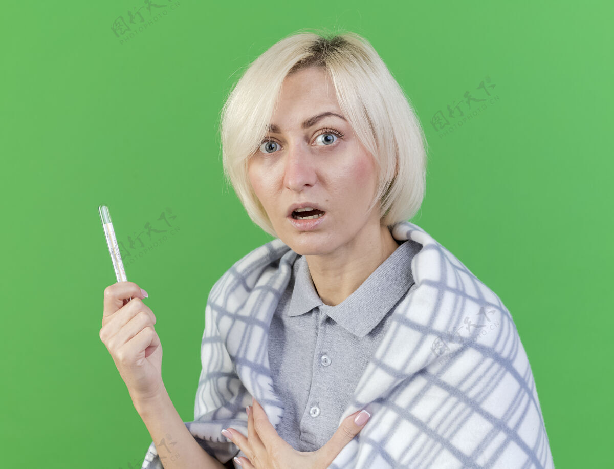 年轻人焦急的年轻金发病女人裹着格子呢拿着温度计看着隔离在绿色墙上的前面焦虑人脸