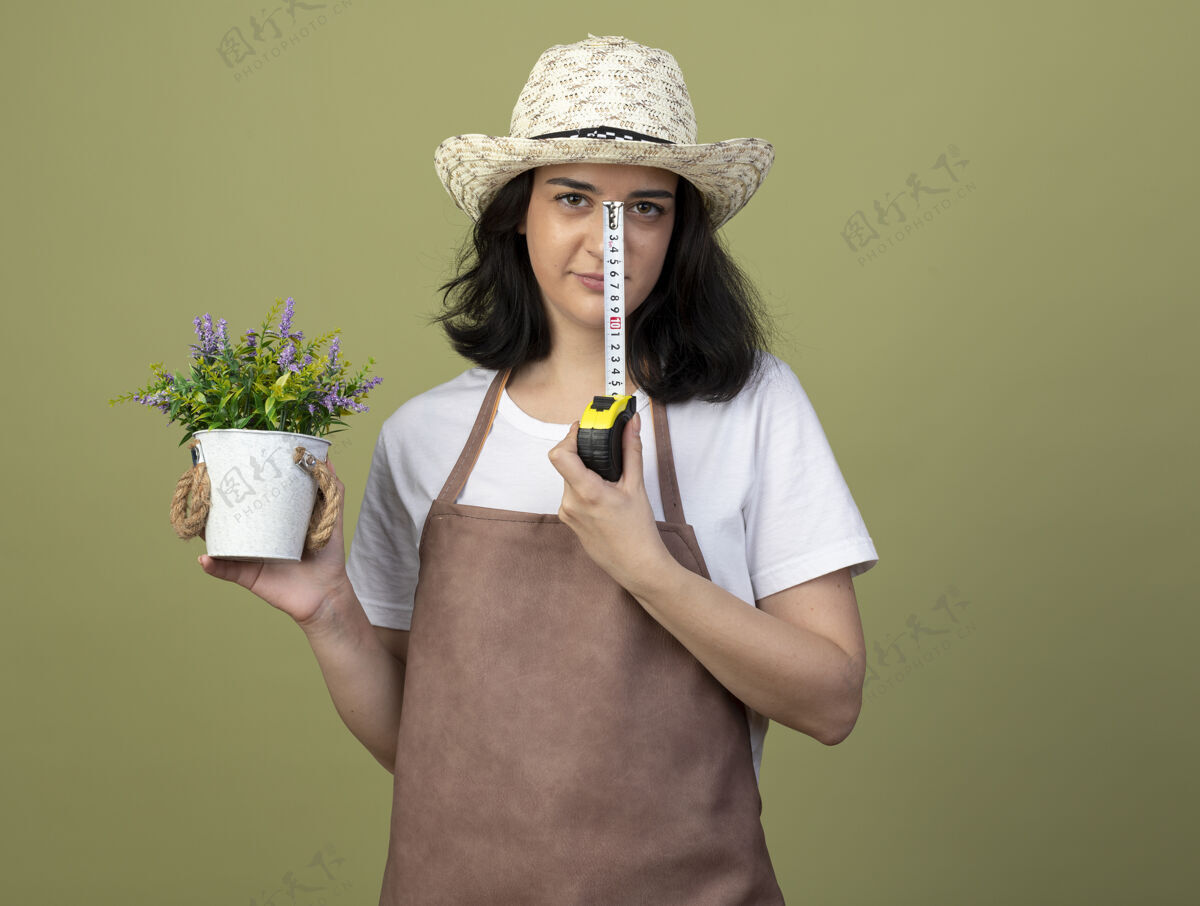 胶带自信的年轻黑发女园丁穿着制服 戴着园艺帽 把花盆和卷尺隔离在橄榄绿的墙上测量表情持有