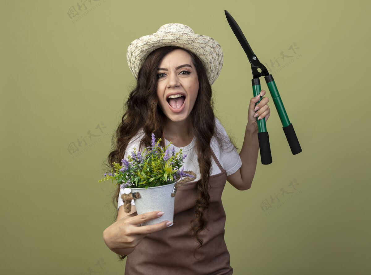 穿身着制服 戴着园艺帽的快乐的年轻女园丁把花盆和园艺剪孤立地放在橄榄绿的墙上橄榄脸制服