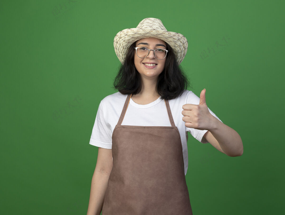 微笑微笑着的年轻黑发女园丁戴着眼镜和制服 戴着园艺帽 在绿色的墙上竖起大拇指拇指姿势人