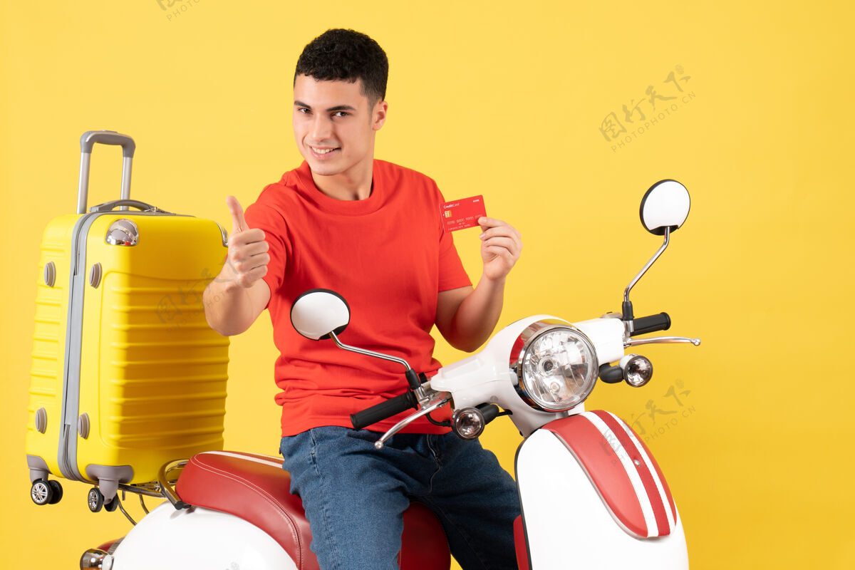 摩托车正面图：坐在轻便摩托车上微笑的年轻人举着信用卡 伸出大拇指前面成人持有