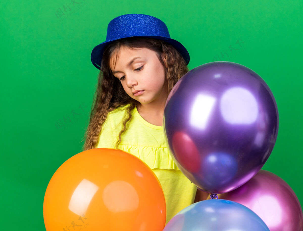 拿着失望的白人小女孩 戴着蓝色派对帽 手里拿着氦气球 孤零零地看着绿色墙壁上的复制品气球派对女孩