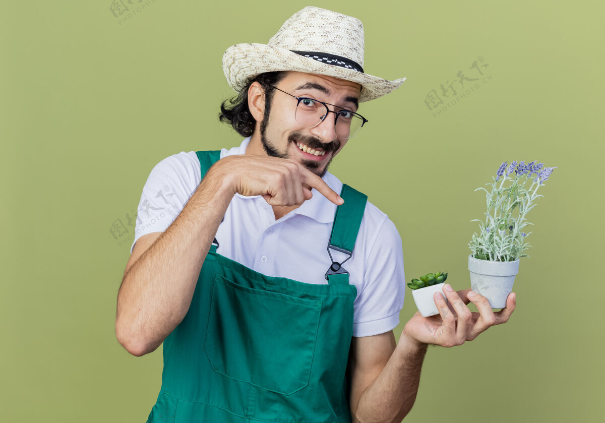 光年轻的留着胡子的园丁 穿着连身衣 戴着帽子 手里拿着盆栽植物 食指指着它 站在浅绿色的墙上欢快地笑着市民目录穿着