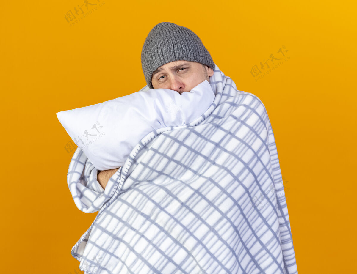 斯拉夫苍白的年轻金发病男子戴着冬天用格子布包裹的帽子抱住隔离在橙色墙上的枕头格子人表情