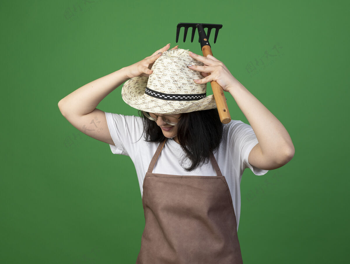 手高兴的是年轻的黑发女园丁戴着眼镜和制服 手上戴着园艺帽 手上拿着耙子隔离在绿色的墙上帽子脸表情