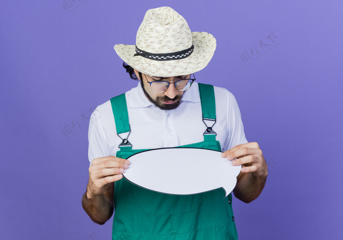 人年轻的留着胡子的园丁 穿着连体衣 戴着帽子 手里拿着一个空白的演讲泡泡牌 严肃的脸站在蓝色的墙上看着它姿势帽子人