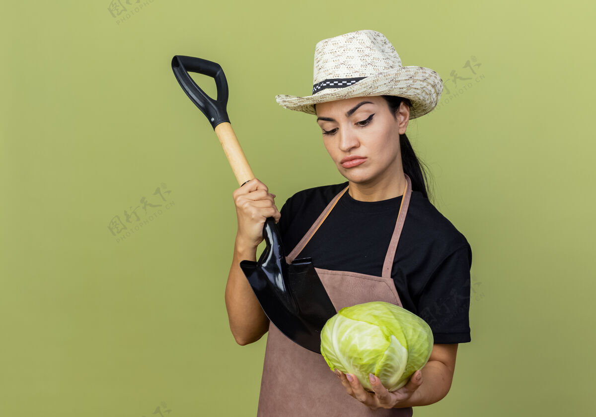 铲子年轻漂亮的女园丁围着围裙 戴着帽子 手里拿着卷心菜和铲子 带着怀疑的表情站在淡绿色的墙上看着卷心菜漂亮衣服站着
